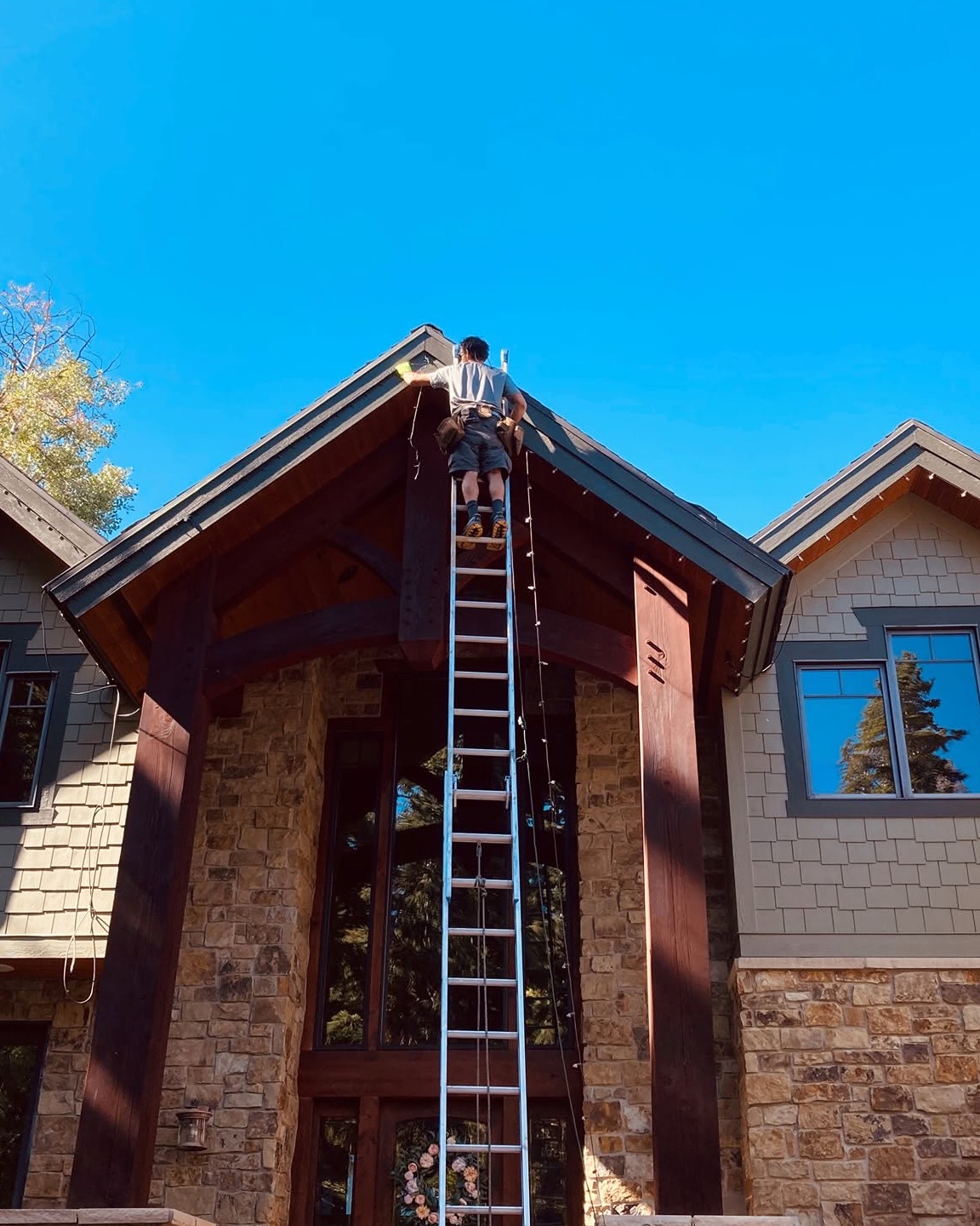 Ryan on a ladder at the peak of a Park City roofline, hanging lights